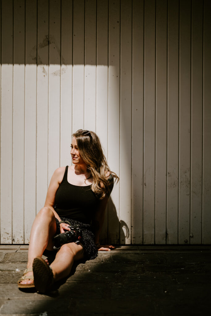 A wedding photographer sits on the ground in front of a sunlit wooden wall, holding a camera in her lap. She wears a black tank top, a patterned skirt, and sandals, with sunglasses resting on her head. She looks to the side and smiles in the warm light.