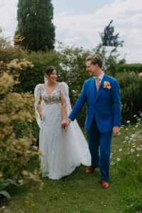 A bride and groom walk hand in hand through a garden, smiling at each other. The bride wears a flowing white gown with floral details and sheer sleeves, while the groom wears a bright blue suit with a pink tie and boutonniere. Lush greenery and flowers surround them under a bright sky.