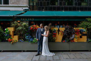 A bride and groom stand outside The Ivy restaurant, smiling and holding each other. Behind them is a large floral display spelling the word "LOVE" in yellow letters decorated with colorful flowers. The groom wears a blue suit, and the bride wears a white sleeveless gown with a train.